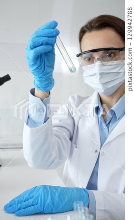 Female scientific researcher holding test tube, analyzing blue liquid while wearing protective gear during medical experiment in sterile laboratory setting. Medicine and science concept Female scientific researcher holding test tube, analyzing blue liquid while wearing protective gear during medical experiment in sterile laboratory setting. Medicine and science concept 129942788
