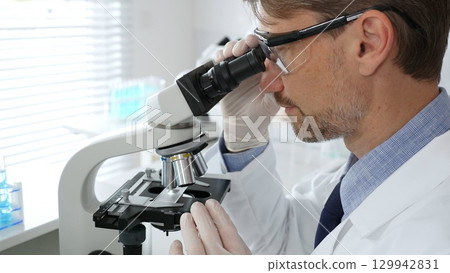 Medical scientist wearing gloves and protective glasses examining samples under a microscope in a bright laboratory filled with test tubes. Medicine, healthcare and science concept Medical scientist wearing gloves and protective glasses examining samples under a microscope in a bright laboratory filled with test tubes. Medicine, healthcare and science concept 129942831