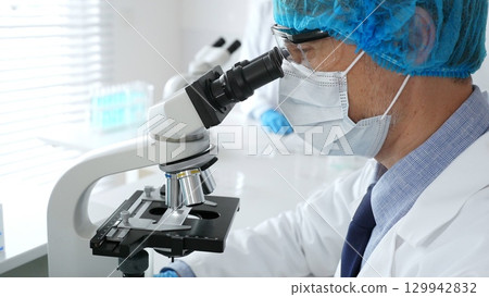 Male scientist in protective mask, cap and gloves studying samples under microscope in sterile laboratory. Concept of medicine, healthcare and science Male scientist in protective mask, cap and gloves studying samples under microscope in sterile laboratory. Concept of medicine, healthcare and science 129942832