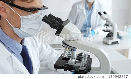 Medical researcher analyzing sample under microscope, wearing protective gear in sterile laboratory setting, performing scientific research. Medicine, healthcare and science concept 129942857