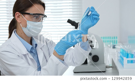 Female scientist wearing a face mask and blue gloves, carefully dropping liquid from a pipette into a test tube in a modern laboratory. Medicine and science concept 129942859