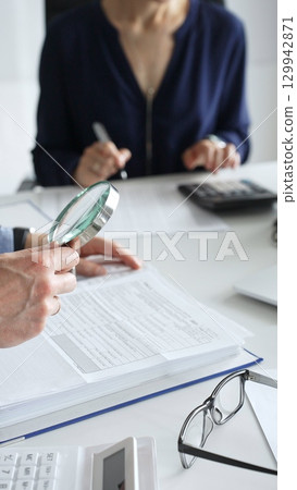 Business colleagues are working together, analyzing financial documents with a magnifying glass, calculating and taking notes in the office. Audit and taxes in business Business colleagues are working together, analyzing financial documents with a magnifying glass, calculating and taking notes in the office. Audit and taxes in business 129942871