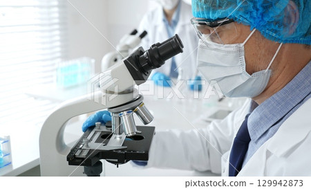 Senior male scientist wearing a protective mask, cap, and gloves, examining samples under a microscope in a modern laboratory. Medicine, healthcare and science concept 129942873