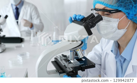 Female scientist wearing a mask and blue safety gear, using a microscope for research while a male colleague works in the background. Medicine, healthcare and science concept 129942877