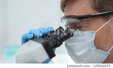 Close up of a scientist researching with a microscope, wearing protective glasses and a mask in a laboratory setting. Medicine, healthcare and science concept Close up of a scientist researching with a microscope, wearing protective glasses and a mask in a laboratory setting. Medicine, healthcare and science concept 129942879