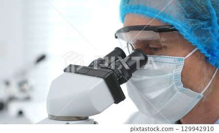 Masked scientist examining sample through microscope, wearing protective lab gear during medical research. Medicine, healthcare and science concept 129942910