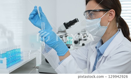 Female scientist wearing a face mask and blue gloves, carefully dropping liquid from a pipette into a test tube in a modern lab. Medicine, healthcare and science concept Female scientist wearing a face mask and blue gloves, carefully dropping liquid from a pipette into a test tube in a modern lab. Medicine, healthcare and science concept 129942926