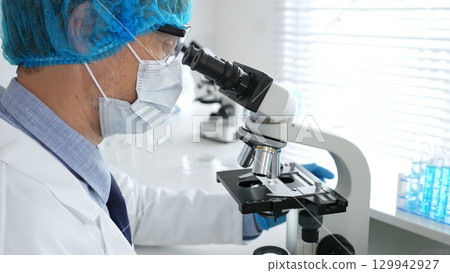 Senior male scientist wearing a protective mask, cap, and gloves, examining samples under a microscope in a modern laboratory. Medicine, healthcare and science concept 129942927