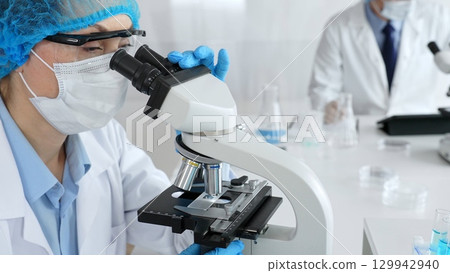 Female scientist wearing a mask and blue safety gear, using a microscope for research while a male colleague works in the background. Medicine, healthcare and science concept Female scientist wearing a mask and blue safety gear, using a microscope for research while a male colleague works in the background. Medicine, healthcare and science concept 129942940