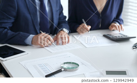 Magnifying glass and calculator over financial documents opposite financial team of male and female auditors, reviewing reports, collaborating on accounting details in office. Audit and taxes 129942949