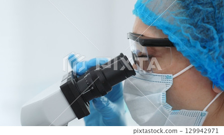 Scientist examining sample under microscope, wearing protective lab gear, performing detailed medical research. Medicine, healthcare and science concept 129942971