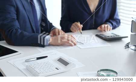 Close up of calculator and magnifying glass over financial documents opposite financial team analyzing data, reviewing reports, collaborating at modern office workspace. Audit and taxes in business 129943000