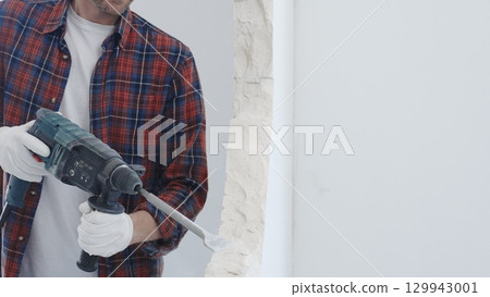 Male construction worker wearing red checkered shirt and protective white gloves is demolishing white wall with rotary hammer drill and generating dust during renovation project, closeup view Male construction worker wearing red checkered shirt and protective white gloves is demolishing white wall with rotary hammer drill and generating dust during renovation project, closeup view 129943001