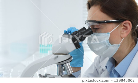 Female scientist wearing a mask and protective glasses using a microscope in a laboratory setting, conducting scientific research and analysis. Medicine, healthcare and science concept Female scientist wearing a mask and protective glasses using a microscope in a laboratory setting, conducting scientific research and analysis. Medicine, healthcare and science concept 129943002