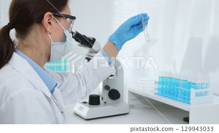 Female researcher wearing protective mask and blue gloves is examining liquid in test tube, sitting near microscopes and lab equipment in laboratory. Medicine, healthcare and science concept 129943033