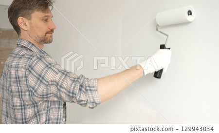 Man professional painter wearing checkered shirt, is rolling white paint on apartment wall with paint roller, transforming interior space with steady strokes during comprehensive home renovation 129943034