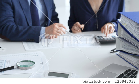 Magnifying glass and calculator over financial documents opposite financial team of male and female auditors, reviewing reports, collaborating on accounting details in office. Audit and taxes Magnifying glass and calculator over financial documents opposite financial team of male and female auditors, reviewing reports, collaborating on accounting details in office. Audit and taxes 129943035