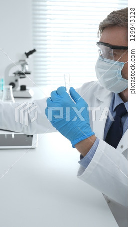 Masked male scientist in lab coat, protective glasses and blue gloves is adding reagent to test tube with microscopes and test tube rack in modern laboratory. Medicine, healthcare and science concept 129943128