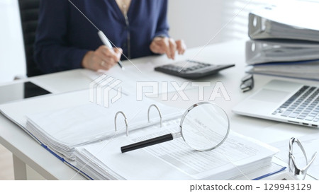 Magnifying glass and calculator are on the table over financial documents opposite female accountant analyzing data, reviewing reports, binders and laptop on a white desk. Audit and taxes concept 129943129