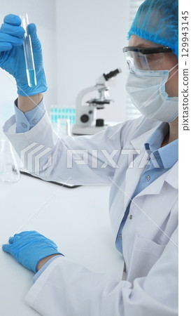 Medical science researcher wearing blue protective gear of cap, gloves and googles, is examining liquid in test tube in laboratory workspace. Medicine and health care concept Medical science researcher wearing blue protective gear of cap, gloves and googles, is examining liquid in test tube in laboratory workspace. Medicine and health care concept 129943145