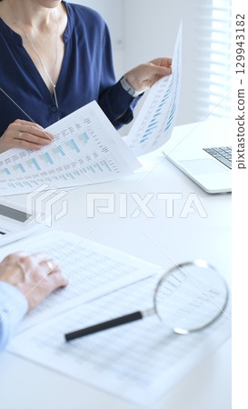 Two professional accountants analyzing financial reports with magnifying glass and calculator, performing accounting and auditing tasks in the office, close up of hands. Audit and taxes in business 129943182