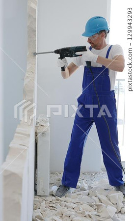 Construction worker demolishing concrete wall with rotary hammer, wearing protective safety gear, generating dust and debris during home renovation work 129943293