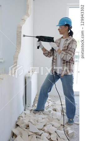 Female construction worker demolishing a wall with a hammer drill, wearing protective gear, generating rubble pile during renovation project 129943302