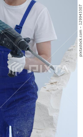 Construction worker demolishing white wall with rotary hammer drill, wearing blue overalls and protective white gloves, generating dust during renovation project, closeup view 129943307