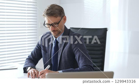 Senior businessman wearing suit, tail and black glasses is concentrating while writing on a document at his desk in a bright, modern office, demonstrating professionalism. Business people concept 129943316