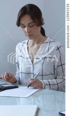 A professional woman calculates finances, checks documents, and accurately and carefully tracks business expenses at a glass table. Audit and taxes in business. 129943339