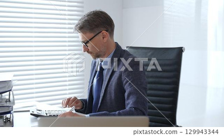 Mature entrepreneur in suit, tie and glasses is sitting at desk, writing notes and using calculator in modern, bright office. Audit and taxes in business 129943344