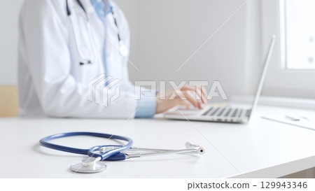 Closeup of blue stethoscope is lying on the table near female doctor working on laptop, highlighting digital tools transforming healthcare diagnostics and patient documentation. Medicine concept 129943346