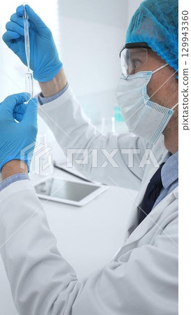 Male medical scientist wearing a lab cap, face mask, googles and blue gloves, carefully dropping liquid from a pipette into a test tube in a modern laboratory setting. Medicine, health care concept 129943360