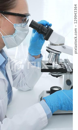 Female scientist wearing blue protective gear analyzing test samples under microscope in laboratory research environment. Medicine, healthcare and science concept 129943364