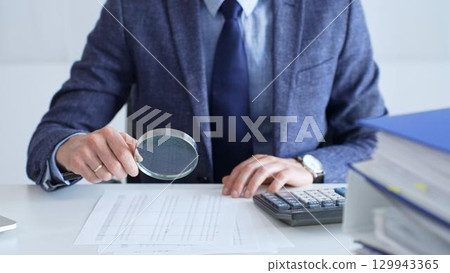 Professional accountant is examining financial documents, magnifying glass in hand, calculator positioned on sleek desk workspace. Audit and taxes in business 129943365
