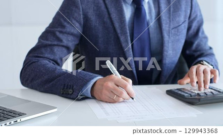 Businessman wearing suit and tie is using calculator and taking notes while working at desk in office, with laptop and stack of folders nearby, close up. Audit and taxes theme in business 129943368