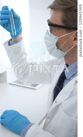 Scientist in a white lab coat, face mask, and gloves, analyzing a blue liquid in a test tube within a laboratory setting. Medicine, healthcare and science concept Scientist in a white lab coat, face mask, and gloves, analyzing a blue liquid in a test tube within a laboratory setting. Medicine, healthcare and science concept 129943369