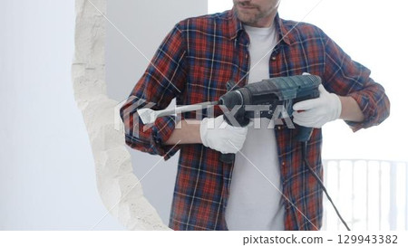 Male construction worker wearing red checkered shirt and protective white gloves is demolishing white wall with rotary hammer drill and generating dust during renovation project, closeup view 129943382