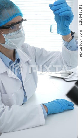 Medical science researcher wearing blue protective gear of cap, gloves and googles, is examining liquid in test tube in laboratory workspace. Medicine and health care concept 129943391