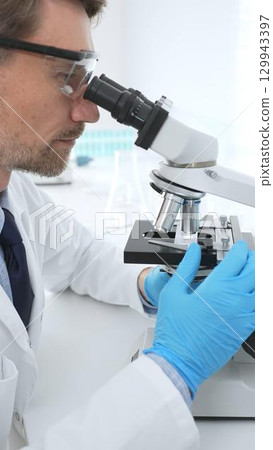 Male medical scientist wearing protective googles and blue gloves, is using microscope analyzing samples in modern laboratory doing scientific research. Medicine, health care and science concept Male medical scientist wearing protective googles and blue gloves, is using microscope analyzing samples in modern laboratory doing scientific research. Medicine, health care and science concept 129943397