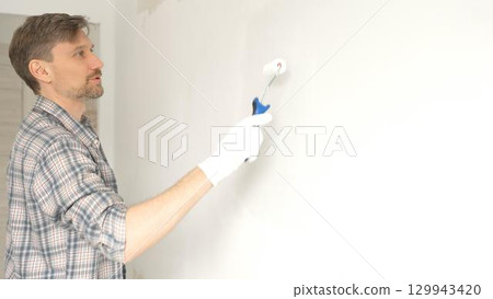 Man professional painter wearing checkered shirt, is rolling white paint on apartment wall with small blue roller, transforming interior space with steady strokes during comprehensive home renovation 129943420