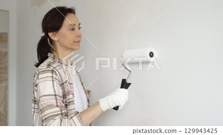 Middle aged woman wearing gloves is skillfully applying paint to a white wall with a paint roller in an apartment undergoing renovation, transforming the space with her creative touch Middle aged woman wearing gloves is skillfully applying paint to a white wall with a paint roller in an apartment undergoing renovation, transforming the space with her creative touch 129943425