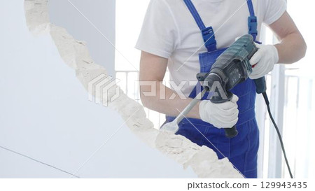 Male construction worker demolishing white wall with rotary hammer drill, wearing blue overalls and protective white gloves, closeup view 129943435