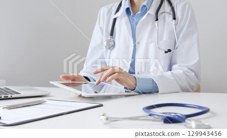 Female doctor wearing white medical coat and stethoscope using digital tablet with laptop and medical records on desk in medical office, showcasing modern healthcare technology. Medicine and helthcare Female doctor wearing white medical coat and stethoscope using digital tablet with laptop and medical records on desk in medical office, showcasing modern healthcare technology. Medicine and helthcare 129943456