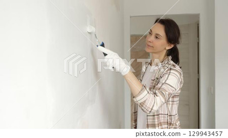 Middle aged woman wearing protective gloves painting white wall with paint roller, updating interior and giving room clean, modern look during home renovation Middle aged woman wearing protective gloves painting white wall with paint roller, updating interior and giving room clean, modern look during home renovation 129943457