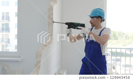 Construction worker demolishing concrete wall with rotary hammer, wearing protective safety gear, generating dust and debris during home renovation work Construction worker demolishing concrete wall with rotary hammer, wearing protective safety gear, generating dust and debris during home renovation work 129943458