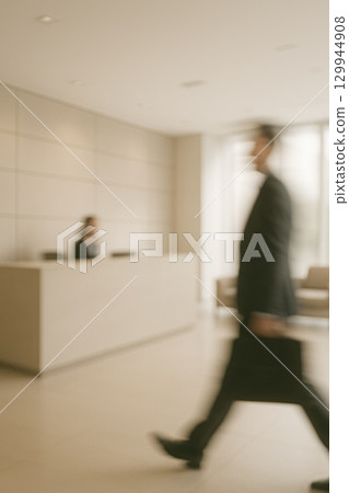 Blurred motion businessman walking in modern office lobby with reception desk and natural light creating dynamic atmosphere 129944908