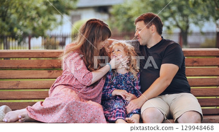 Family bonding on wooden bench, parents hugging young daughter during relaxed backyard moment of connection and love 129948468