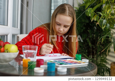Portrait of young girl in red warm sweater drawing in a sketchbook while sitting on the balcony 129949112