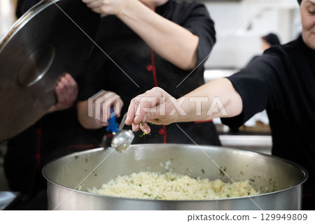 Group of chefs in black uniforms preparing rice in a large pot in a commercial kitchen. Female cooks stirring and adding herbs while working as a team. Concept of teamwork, gastronomy, and hospitality Group of chefs in black uniforms preparing rice in a large pot in a commercial kitchen. Female cooks stirring and adding herbs while working as a team. Concept of teamwork, gastronomy, and hospitality 129949809